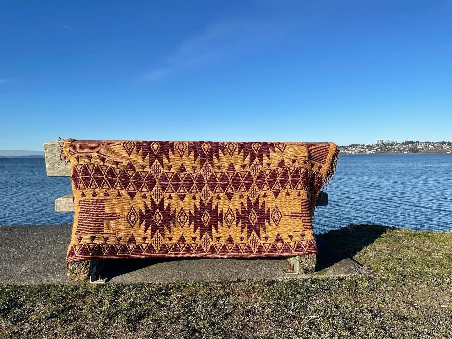 A burgundy and ochre geometric crochet blanket draped over a driftwood bench by the sea.